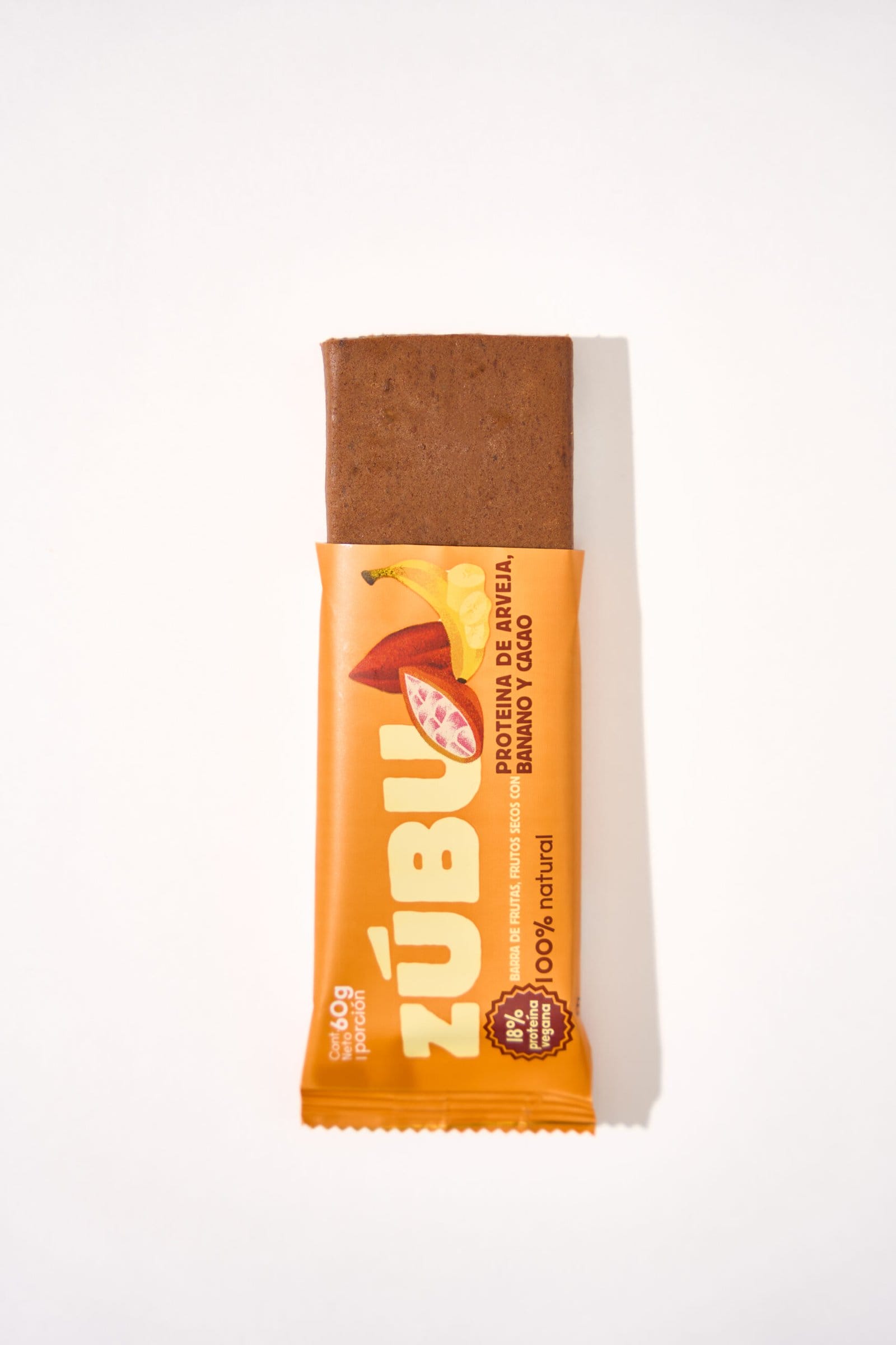 Una barrita de proteínas ZUBU "con arveja, chocolate 70% y coco" en envase naranja con ilustraciones de plátano, arveja y cacao sobre fondo blanco.